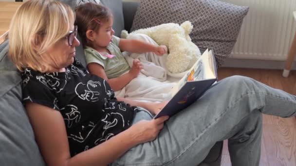 Γιαγιά με το παιδί ανάγνωση βιβλίο στον καναπέ στο σαλόνι. Οικογενειακός χρόνος δέσμευσης — Αρχείο Βίντεο
