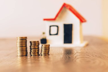 Paralar yığılmış ve arka plan üzerinde bulanık bir ev ile bir ahşap masa üstünde: Emlak, gayrimenkul yatırım, evi ipotek, tasarruf mali kavramı.