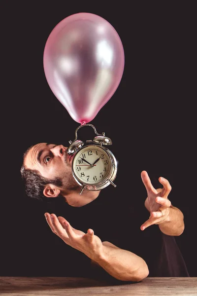 Time flies concept - picture of a man in panic trying to grab an old metal clock that is attached to a pink balloon flying above a wooden table.