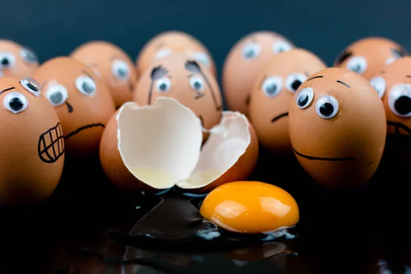 Fun concept: raw eggs with googly eyes and drawn features are in shock and sad as they witness another egg broken in two with the white and yolk in front of them; dark background.