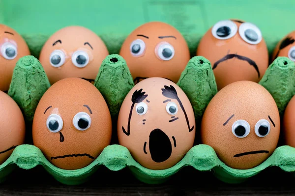 Fun concept: raw eggs with googly eyes and drawn features are in shock and sad while they sit in a green carton box on top of a wooden table
