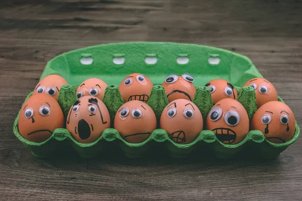 Fun concept: raw eggs with googly eyes and drawn features are in shock and sad while they sit in a green carton box on top of a wooden table