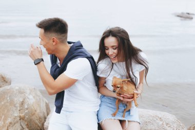 Genç güzel pozitif çift ile deniz taş üzerinde oturan küçük köpek. Yaşam tarzı ve Evcil hayvan