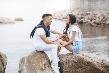 Genç güzel pozitif çift ile deniz taş üzerinde oturan küçük köpek. Yaşam tarzı ve Evcil hayvan