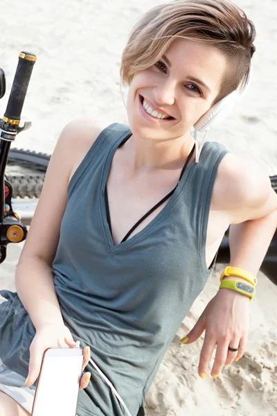 Young smiling active woman sits on sand with bicycle and phone.  Lifestyle, positive and active