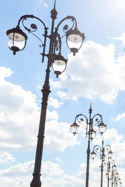 Row of graceful triple street lamp posts under sunlight against blue sky with clouds. Urban decor and electrical equipment