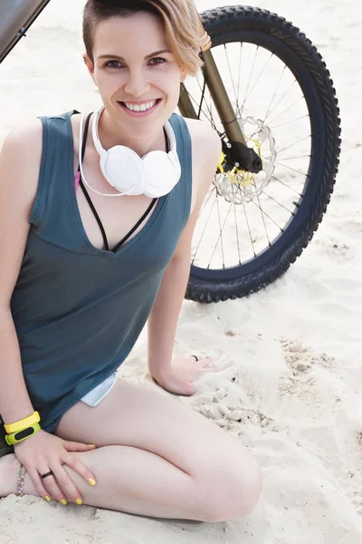 Young smiling active woman sits on sand with bicycle
