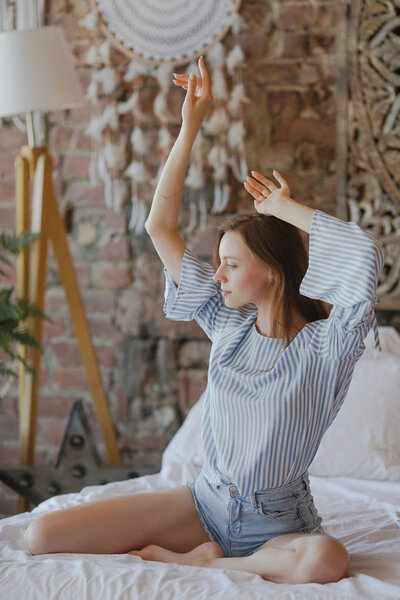 Молодая милая девочка сидит на кровати и протягивает руки вверх
