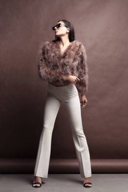 Tüylü pembe pelerinli, yapay kürklü ve kahverengi arka planda bej renkli pantolonu olan zarif genç bir kız. Moda retro tarzı