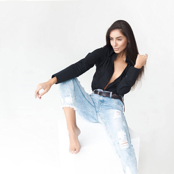 Молодая крутая хипстерша в черной рубашке и синие банановые джинсы девушка сидит на белом кубе на белом фоне. Модный современный стиль
