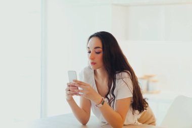 Ciddi, genç ve güzel bir kadın minimalist hafif mutfakta telefonu kullanıyor. İçerik oluşturma ve iletişim. Arama ya da mesaj bekleniyor