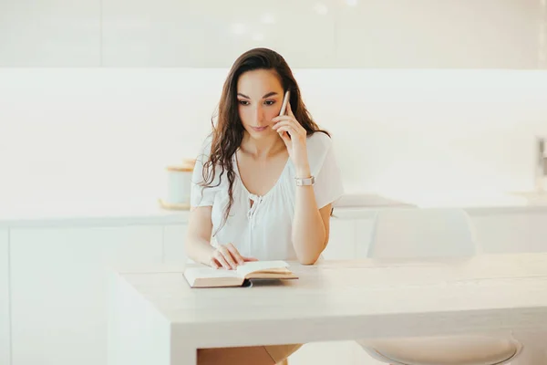 Serious beautiful woman speaks on phone in minimalistic light kitchen. Technology and leisure. Communication and emotions at home