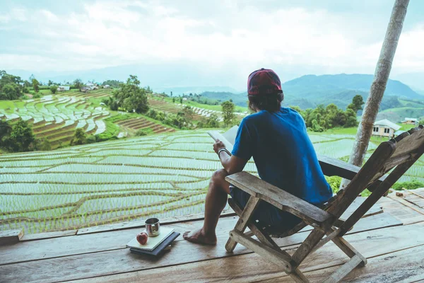 Asian man travel nature. Travel relax. sit reading book the balcony of the resort. View of the field on the Moutain. laptop, computer, education, read a book, book, nature, outdoor,Rice fields, holiday, relax, travel Thailand, summer.