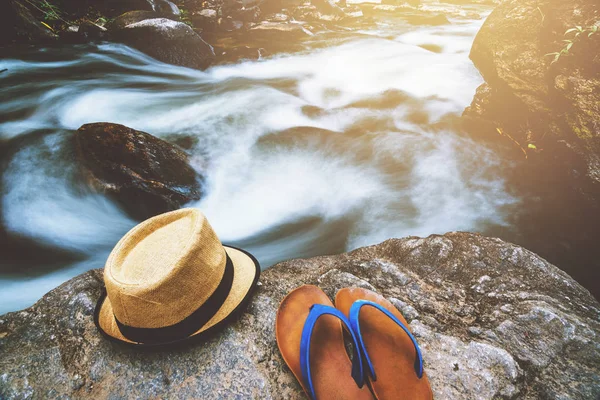 Viaggio natura, Viaggi relax, Cappello con sandali appoggiati su rocce alla cascata . — Foto Stock