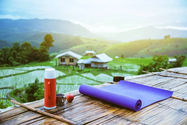 Violette Yogamatte und rote Wasserflasche mit einem Glas Wasser aus rostfreiem Edelstahl vorgestreckt. und roter Apfel auf hölzernem Hintergrund. auf dem Balkon Landschaft natürliche Feld.Reisen entspannen. papongpieng in thailand — Stockfoto
