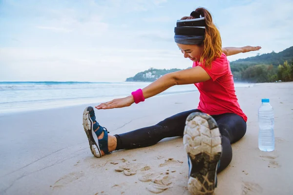 Ασιατικές γυναίκες τζόκινγκ προπόνηση στην παραλία. Καθίστε στο γυμναστήριο της παραλίας χαλαρώστε με τεντωμένα πόδια και τεντώστε το βραχίονα. Τζόκινγκ στην παραλία. ταξίδια στην Ταϊλάνδη, άσκηση. — Φωτογραφία Αρχείου