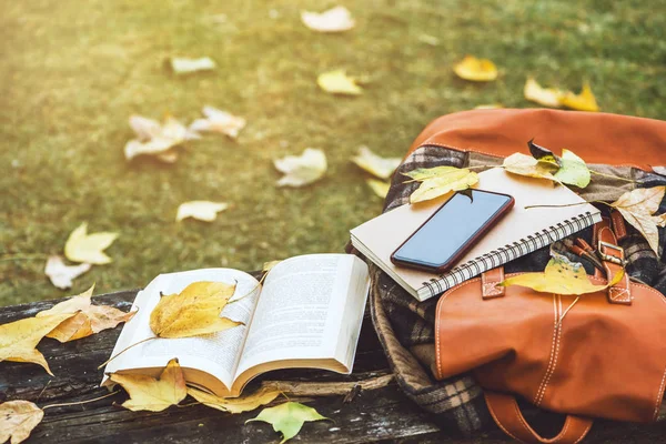La mochila se coloca en una mesa de madera antigua con libros, cuadernos, teléfonos y la naturaleza de las hojas de arce desde la vista superior . — Foto de Stock
