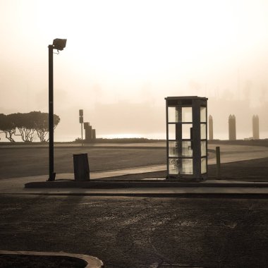An empty parking lot with a street lamp, phone booth and trash can on a foggy morning with water and shadows in the background clipart