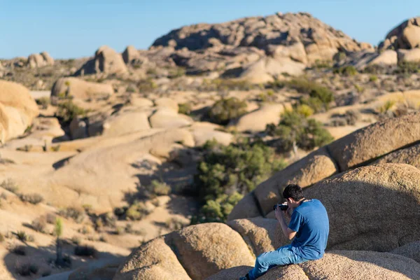在约书亚树国家公园的一个阳光明媚的日子里 一个年轻人坐在岩石 高山和植物的背景下 用相机拍照 — 图库照片