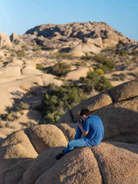 若い男は ジョシュアツリー国立公園で晴れた日に背景に岩 山や植物と砂漠の岩の上に座っている間 カメラで写真を撮ります — ストック写真
