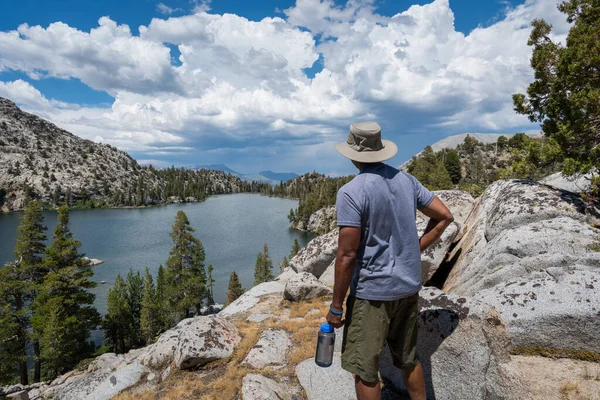 An African American hiker resting at a higher elevation, taking in the views of the lake, trees, mountains and clouds in the distance in California.