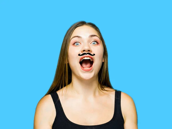 Young caucasian woman with drawn moustache on her face, screaming opening the mouth; indoors, isolated, studio, blue background