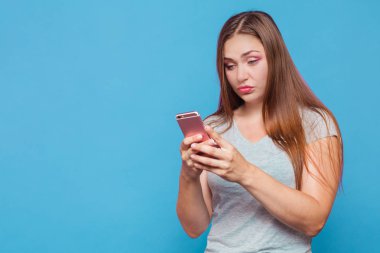 Kahverengi saçlı, mavi gözlü seyir için pembe telefon inanmaz yüz buruşturma ile Ördeksurat Genç beyaz kadınla. Giyiyor rahat (gri bluz). Mavi arka plan, stüdyo, izole, modeli, kopya alanı.