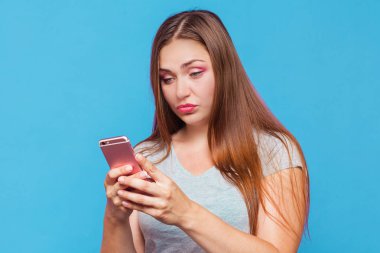 Kahverengi saçlı, mavi gözlü seyir için pembe telefon inanmaz yüz buruşturma ile Ördeksurat Genç beyaz kadınla. Mavi arka plan, stüdyo, izole, modeli, kopya alanı, kavram.