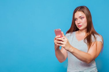 Kahverengi saçlı, mavi gözlü seyir için pembe telefon şaşkınlık yüz buruşturma ve hafif slmile ile genç beyaz kadınla. Mavi arka plan, stüdyo, izole, kopya alanı, modeli.