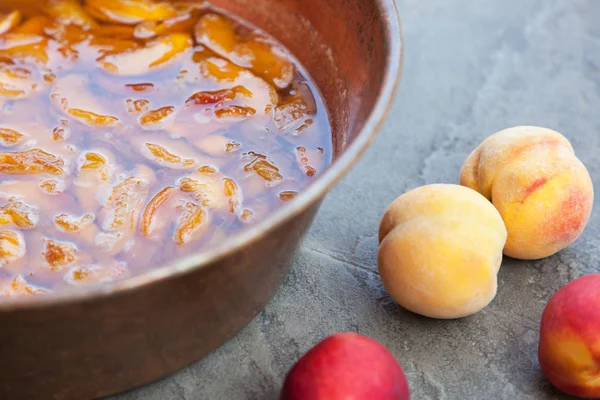 Fresh bright peaches and nectarins lay on the stone bricks, close to the copper jar, full of peach jam. Close up, outdoor, copy space. Fun of cooking.
