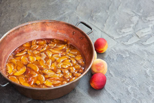 Fresh bright peaches and nectarins lay on the stone bricks, close to the copper jar, full of peach jam. Close up, outdoor, copy space. Fun of cooking.