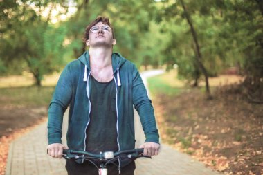 Romantik Genç beyaz yakışıklı erkek kıvırcık saçlı yalnız yeşil park yol bisiklet kadar görünüyor. Yuvarlak gözlük altın küpe bir kulakta, raslantısal spor giyim. Sağlıklı yaşam kavramı, açık havada, kopya alanı. Akıllı göz.