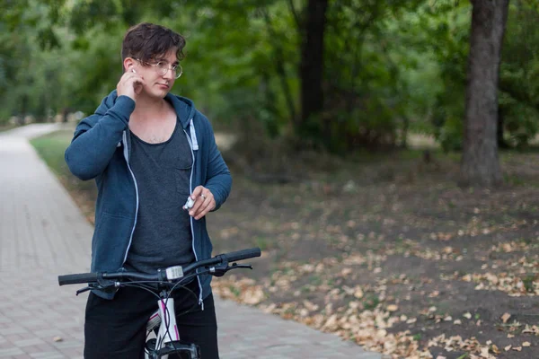Attractive young caucasian man with dark hair bicycling in the park. Touches the white earphone (airpods). Outdoors, autumnl day. Diversity people. Melancholy mood. Round golden glasses, silver ring on one finger, white and pink bicycle. Copy spase