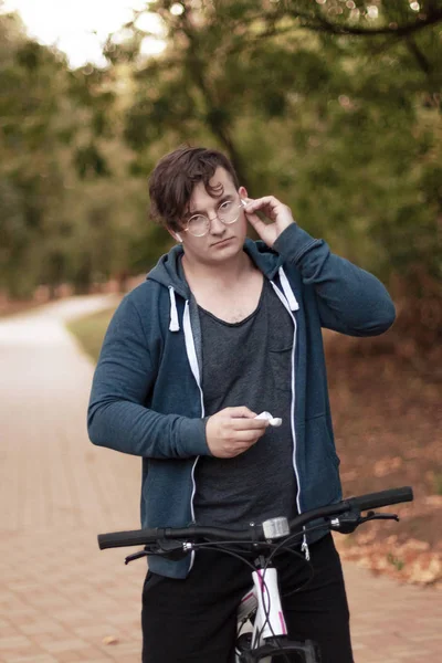 Attractive young caucasian man with dark hair bicycling in the park. Touches the white earphone (airpod). Outdoors, autumn day. Diversity people. Melancholy mood. Round golden glasses, silver ring on one finger. Copy space. Favorite music and autumn