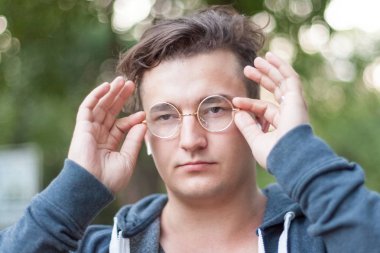 Koyu saçlı çekici genç bir beyaz adam portresi yakından yuvarlak altın gözlüğü burnuna tutar. Ciddi ifade, duygular gözünde. Açık havada, yeşil arka plan. Çeşitlilik insanlar. Beyaz kulaklık (airpods), gümüş yüzük parmağına. 