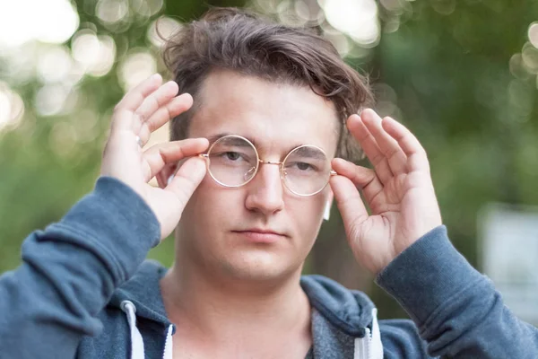 Close up portrait of attractive young caucasian man with dark hair holds his round golden glasses on the nose. Serious expression, emotions in the eyes. Outdoors, green background. Diversity people. White earphones (airpods), silver ring on finger.