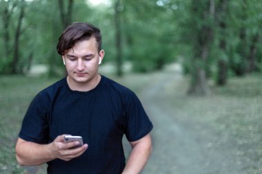 Yeşil terk edilmiş park telefon ekranında çekici beyaz delikanlı bakar. Beyaz küpe, rahat kıyafet (siyah gömlek), siyah saçlı. Açık havada, portre, kopya alanı kapatın. Yalnızlık ve romantik ruh hali. Arkadaş iletiye kaydediyor. Çeşitlilik