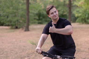 Beyaz yakışıklı genç başparmak yukarıya vasıl belgili tanımlık park bisikleti. Beyaz kulaklık, karanlık rahat kıyafet. Açık havada, terk edilmiş park. Yakın çekim, alanı Kopyala. Gülüşü, duygular ve ifade. Çeşitlilik insanlar. 