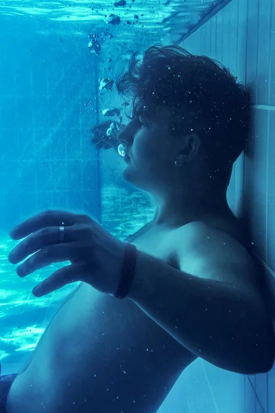 Young attractive caucasian man slowly falls down on the pull floor, underwater. Blue and deep aquamarine background, melancholy atmosphere. Ring and bangle (bracelet) on the hand, earring. Bubbles around. Copy space