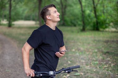 Çekici genç beyaz adam cep telefonu ve bisiklet yeşil parkta tutar. Beyaz kulaklık, küpe, yüzük, karanlık giyim, koyu saç ve gözler. Terk edilmiş park arka plan. Açık havada, yakın çekim, alanı Kopyala. Sabah fitness olumlu duygular ile.