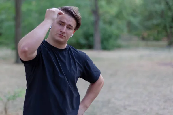 Young handsome caucasian man touches the hair, green abandoned park background. White earphones, ring, dark casual t-shirt, dark hair and eyes. Outdoors, close up, copy space. Model expression and emotions on the face.