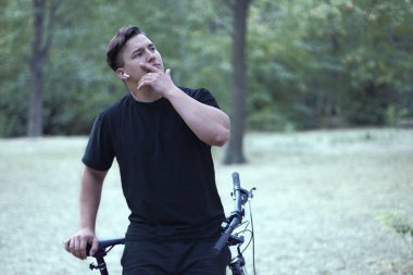 Genç yakışıklı beyaz adam bir modeli stil yüzüne dokunmak onun bisiklet ile duruyor. Beyaz kulaklık, küpe, yüzük, karanlık rahat kıyafet, siyah saçlı. Terk edilmiş park arka plan olarak. Açık havada, kopya alanı.