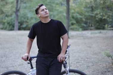 Genç yakışıklı beyaz adam onun bisiklet modeli tarzı ile duruyor. Beyaz kulaklık, küpe, yüzük, karanlık rahat kıyafet, siyah saçlı. Terk edilmiş park arka plan olarak. Açık havada, kopya alanı.