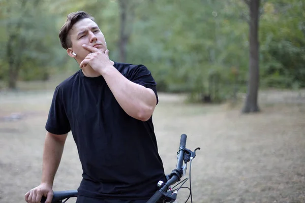 Young handsome caucasian man stands with a bicycle touching his chin in a model style. White earphones, ring, black casual wear, dark hair. Abandoned park as background. Outdoors, copy space.
