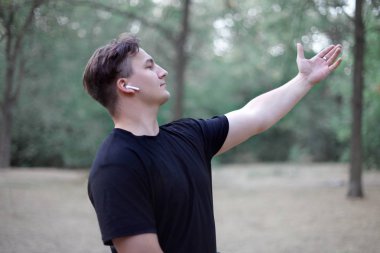 Genç yakışıklı beyaz adam elini devam et, park veya ormanın ortasında uzanan duruyor. Çok ciddi ve Roma kasalı thoughful. Beyaz kulaklık, ring, karanlık rahat kıyafet, siyah saçlı. Terk edilmiş park arka plan olarak. Açık havada, kopya alanı.