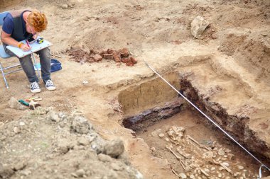 Arkeolojik kazılar. Genç şık arkeolog drowings insan kemik, iskelet ve zemin mezar kafatasında yapar. Gerçek kazıcı işlemi. Kulaklık, cep telefonu, güneş gözlüğü. Araçlar (mezura, rulet, fırça, bıçak). Açık havada, c