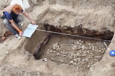 Arkeolojik kazılar. Kızıl saçlı genç şık arkeolog drowings insan kemik, iskelet ve zemin mezar kafatasında yapar. Gerçek kazıcı işlemi. Kulaklık, cep telefonu, güneş gözlüğü. Araçlar (mezura, rulet, fırça, bıçak
