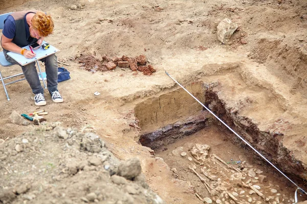 Arkeolojik kazılar. Genç şık arkeolog drowings insan kemik, iskelet ve zemin mezar kafatasında yapar. Gerçek kazıcı işlemi. Kulaklık, cep telefonu, güneş gözlüğü. Araçlar (mezura, rulet, fırça, bıçak). Açık havada, c
