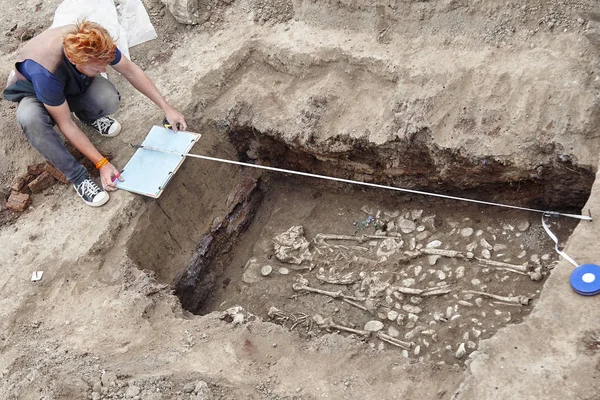 考古发掘 年轻的红头发的时尚考古学家使人类的骨骼 骨骼和头骨淹没在地下墓穴中 真正的挖掘机过程 带耳机 太阳镜 轮盘赌 — 图库照片
