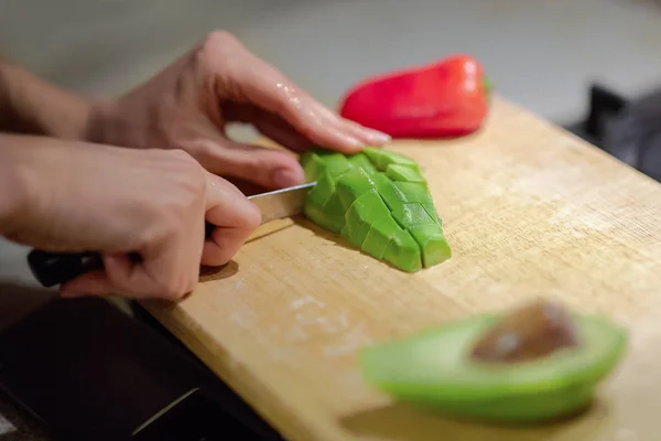 Female hands cutting fresh organic avocado with knife on wooden board in kitchen. Red bell pepper on background. Close up, indoors, copy space.